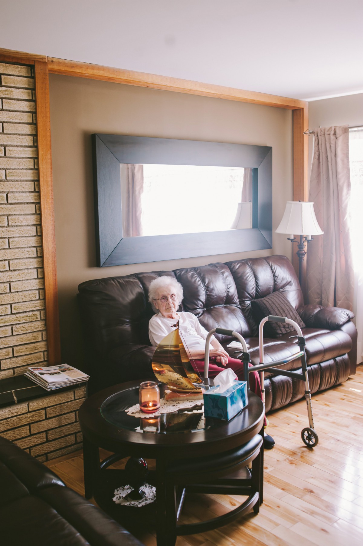 An elderly woman sitting on a sofa in a cozy living room with a walker nearby.