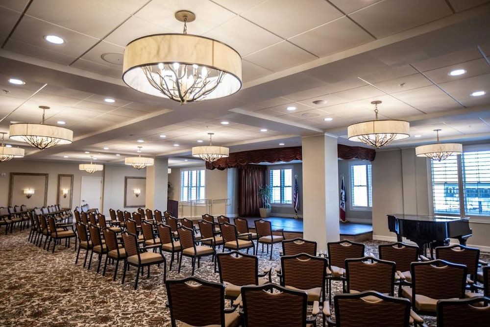 An elegant conference room with arranged seating and a stage, illuminated by chandeliers.