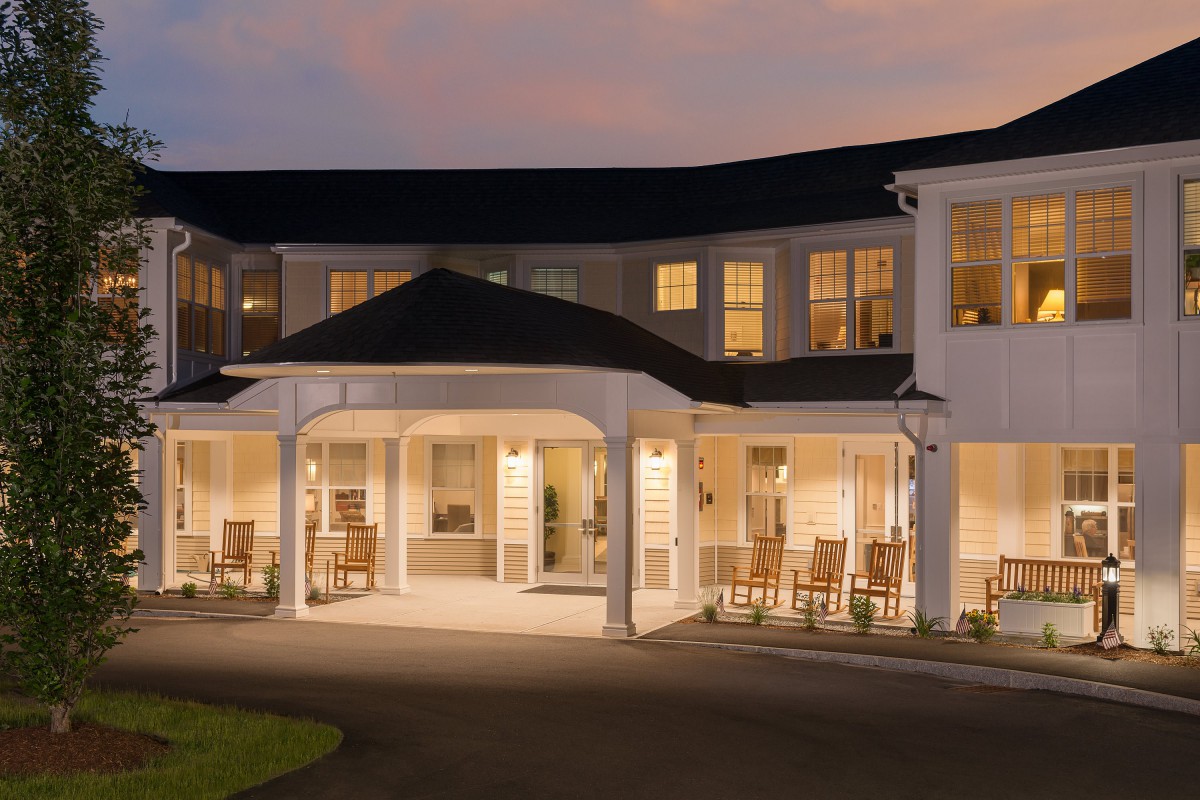 A well-lit building with a porch and rocking chairs at dusk.