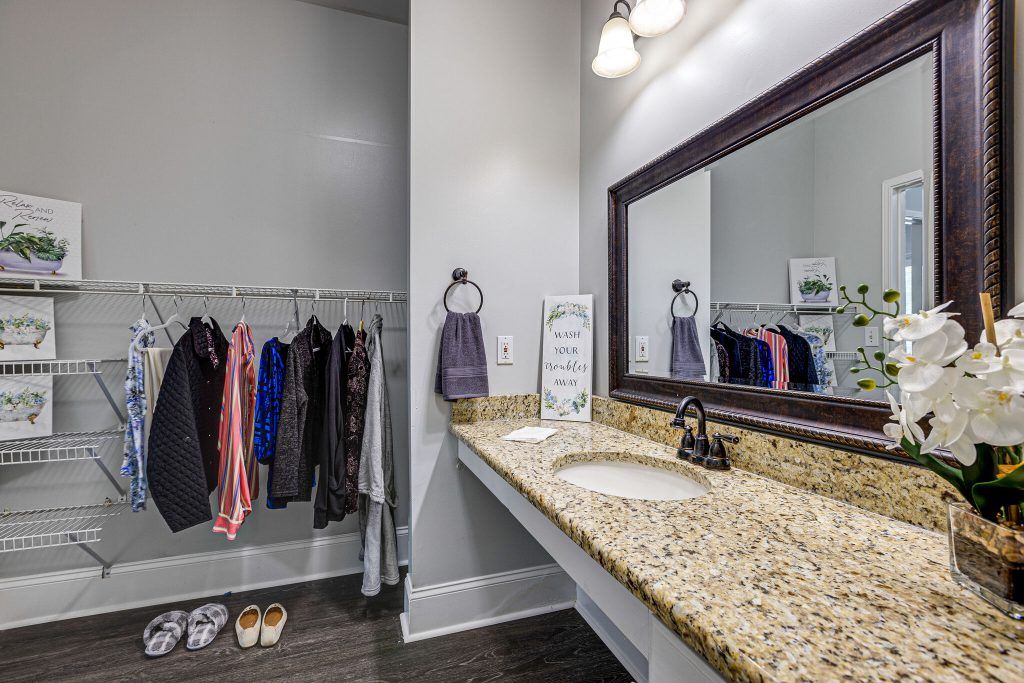 A modern bathroom with a granite countertop, sink, and mirror alongside a clothes rack and lighting fixture.