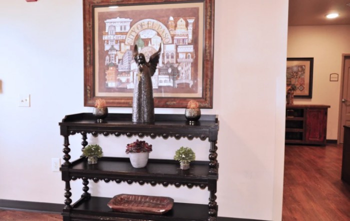 A decorative shelf with a statue, candles, and small plants, set against a wall featuring framed artwork.