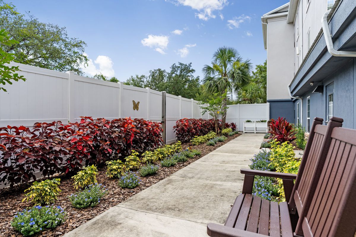 A well-maintained garden with a concrete pathway, colorful flowers, and shrubs alongside a white fence.