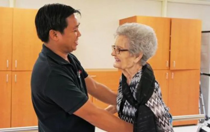 A man and an elderly woman share a warm interaction indoors, smiling at each other.