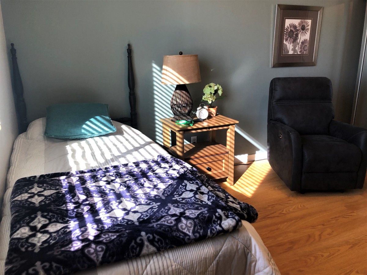 A cozy bedroom with sunlight streaming in, highlighting a bed and a side table with a lamp.