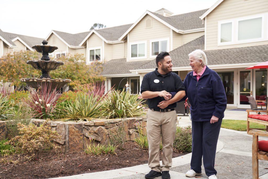 A caregiver assisting a senior citizen as they walk outdoors in a landscaped area.