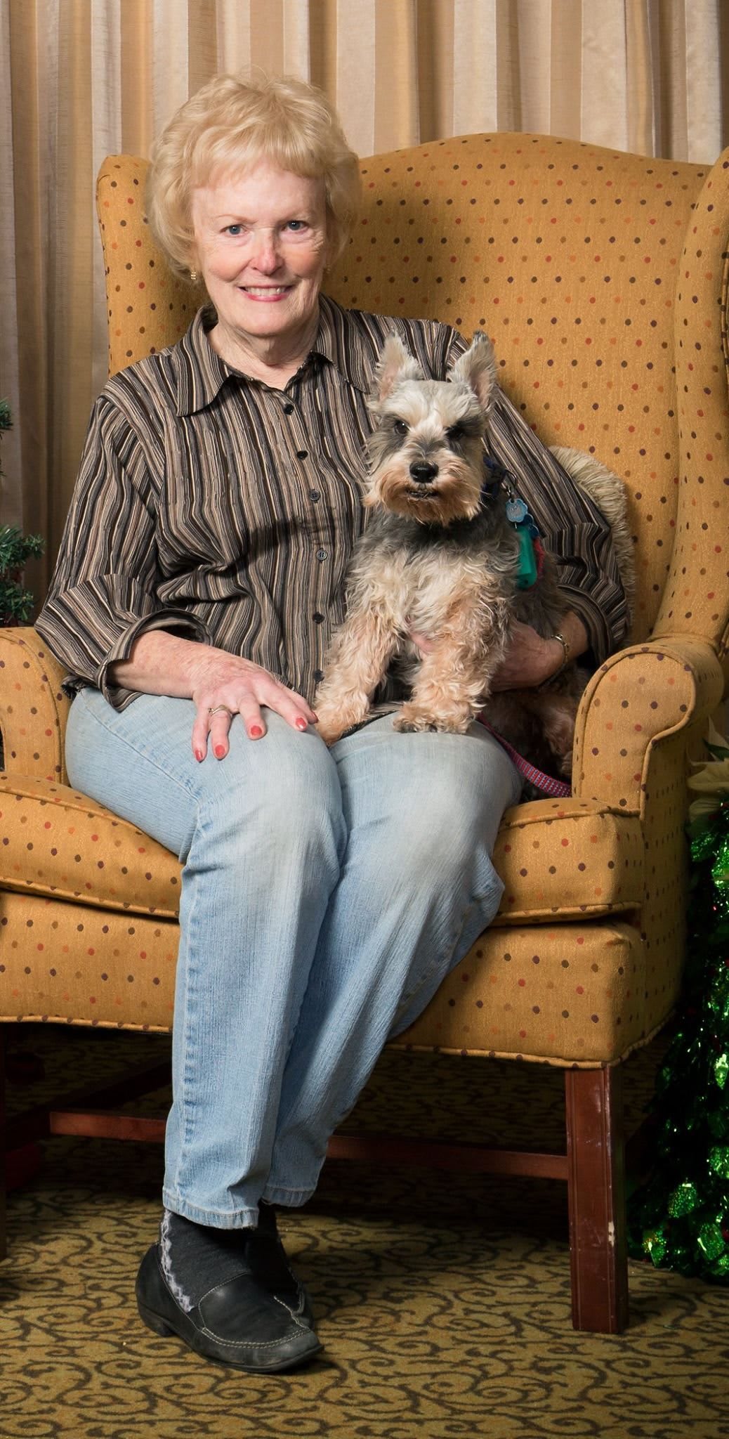 A person sitting indoors on a chair with a small dog on their lap.
