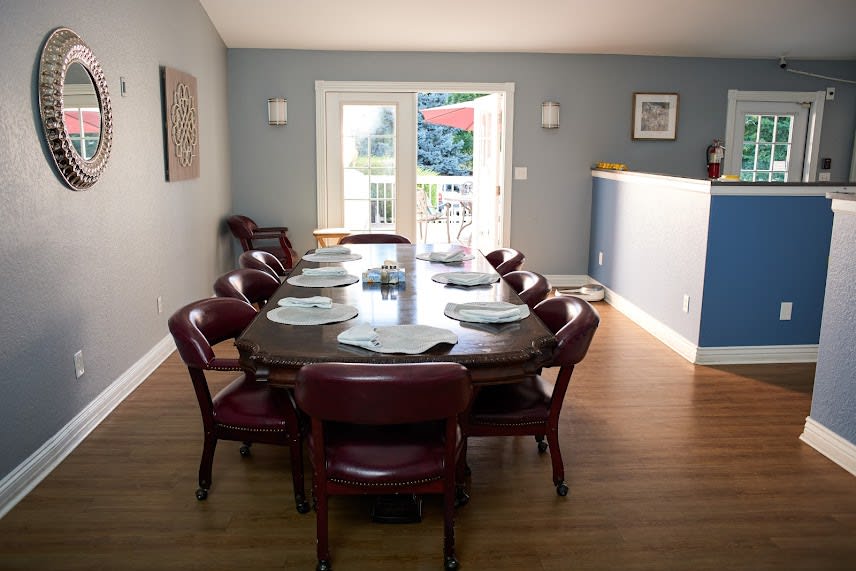 A dining room featuring a long table with place settings, surrounded by chairs and illuminated by natural light from large windows.