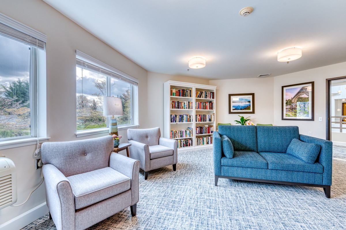 A cozy living room featuring a blue sofa, two gray armchairs, a well-stocked bookshelf, and natural light from a large window.