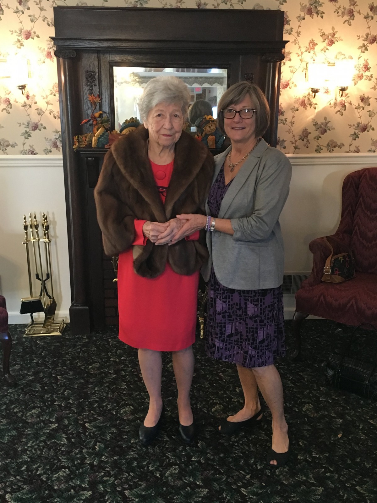 Two women, dressed formally, stand together in a living room setting.
