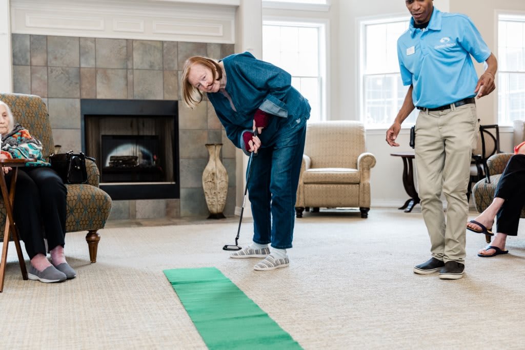 An elderly woman practices putting on an indoor golf green with assistance in a living room setting.