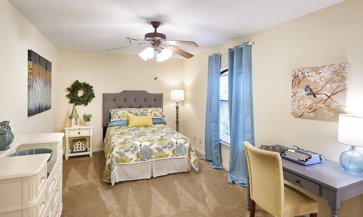 A cozy bedroom with a bed, desk, and ceiling fan, featuring blue curtains and elegant decor.