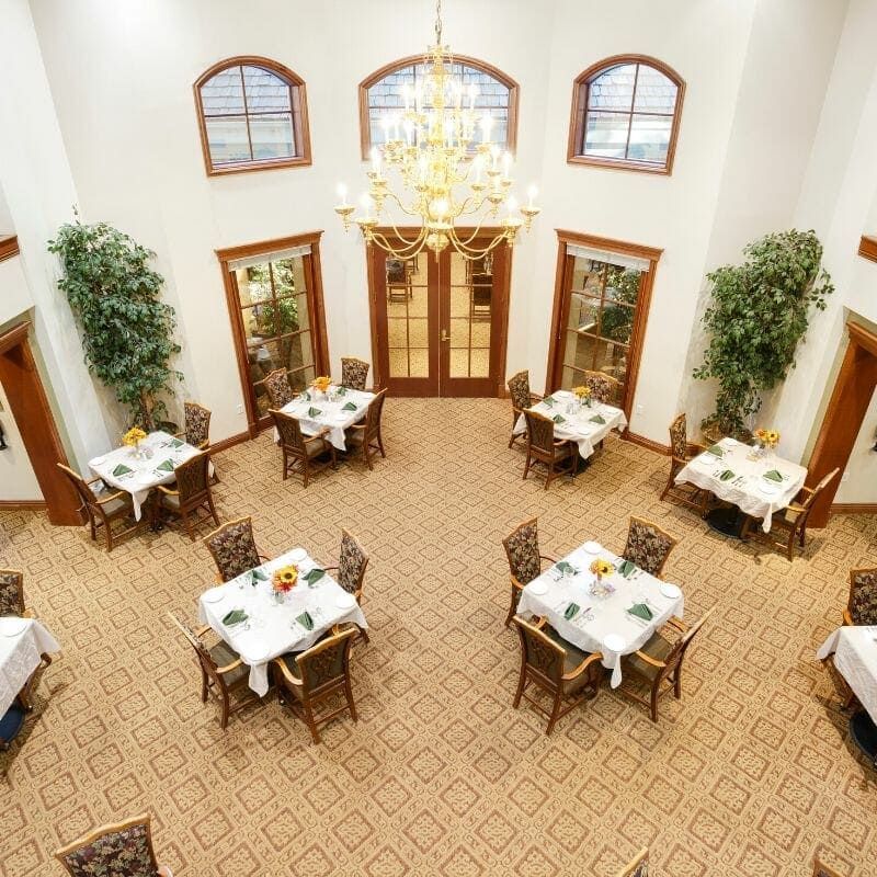An elegant dining room interior with tables set for guests, featuring a chandelier and decorative plants.
