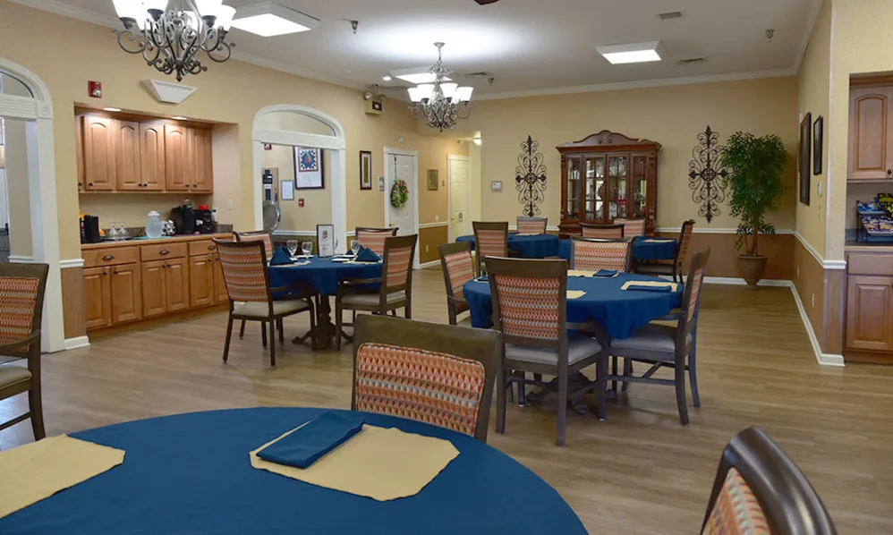 A spacious dining room with multiple tables covered in blue tablecloths, surrounded by wooden chairs and decorated with chandeliers.