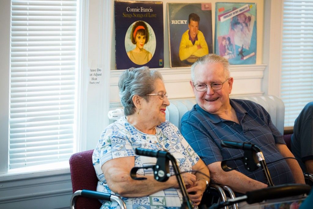 An elderly couple smiling at each other while sitting indoors, surrounded by music album covers.