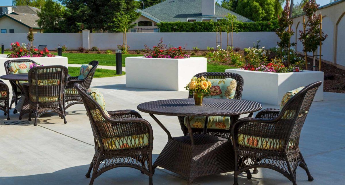 A sunny patio with wicker chairs and a table adorned with flowers, surrounded by a well-maintained garden.