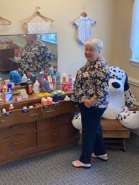 A woman stands beside a dresser filled with toiletries, reflected in the mirror, with a large stuffed animal in the background.