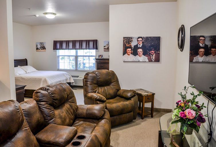 A cozy living space featuring recliner chairs, wall art, and a window with curtains, leading into a bedroom area.