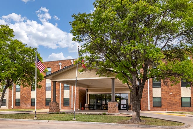 A hotel entrance with flags and a large tree under a partly cloudy sky.