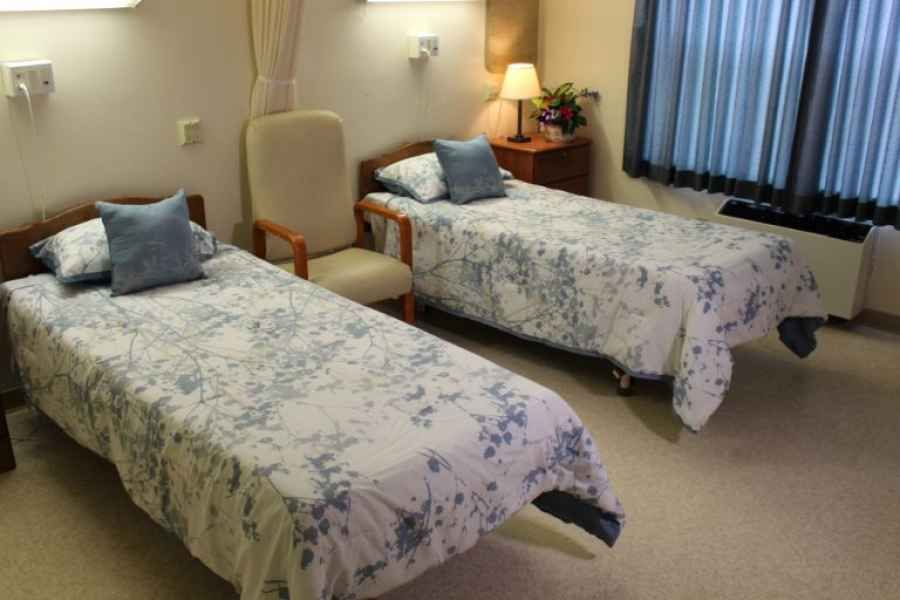 A cozy bedroom with two single beds, each adorned with floral-patterned bedding. A wooden nightstand with a lamp sits between them, alongside a comfortable chair.