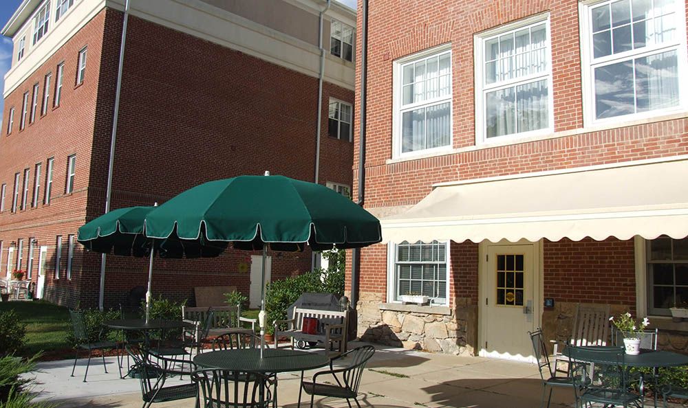An outdoor patio area with tables and chairs under a green umbrella, adjacent to a brick building.
