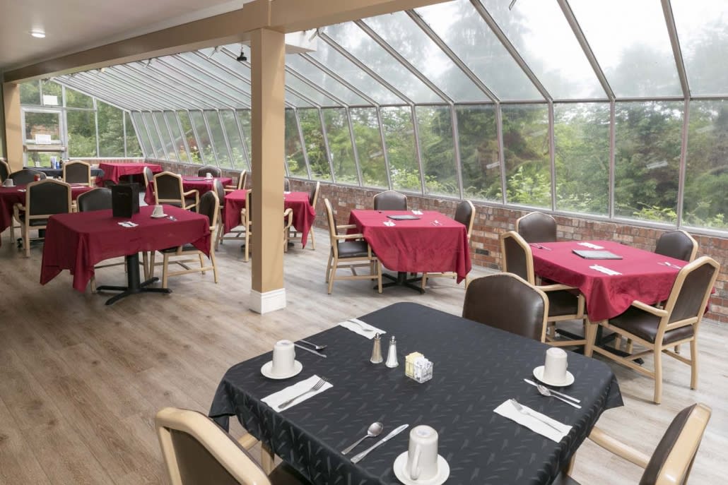 A well-lit restaurant dining area with tables covered in red and black tablecloths, surrounded by large windows and a glass roof.