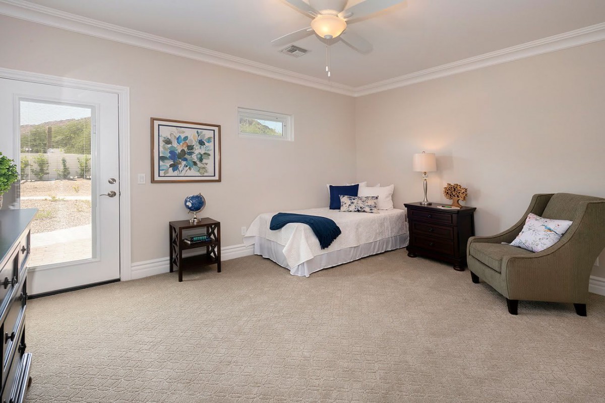 A spacious, neutral-colored bedroom featuring a single bed with blue accents, a nightstand with a lamp, a decorative painting, and a comfortable chair.