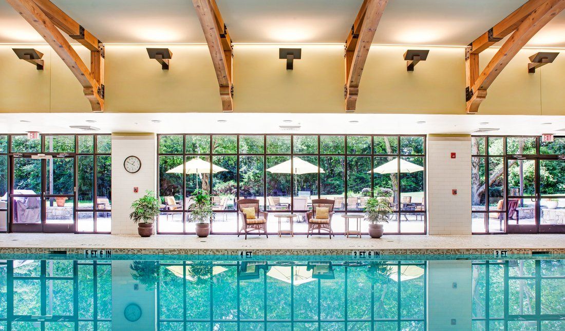 A serene indoor swimming pool area with large glass windows, sun loungers, and umbrellas, surrounded by greenery.