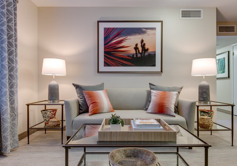 A cozy living room with a stylish gray sofa, accented by orange pillows and framed artwork above. Two matching lamps flank the sofa, providing warm lighting. A glass coffee table sits in front of the sofa, completing the contemporary design.