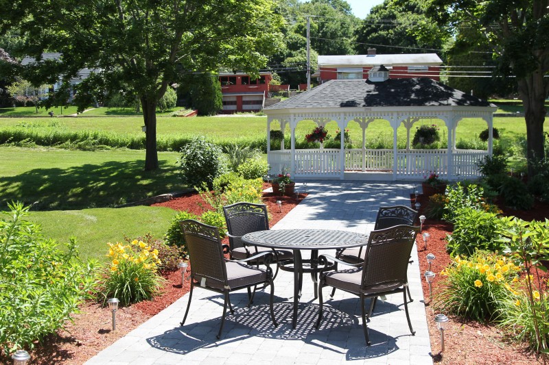 A sunny garden with a patio set and gazebo surrounded by lush greenery.