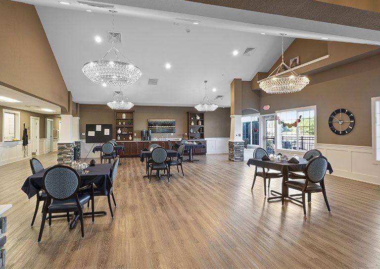 A spacious dining area with elegant chandeliers, multiple tables and chairs, and a kitchenette in the background.