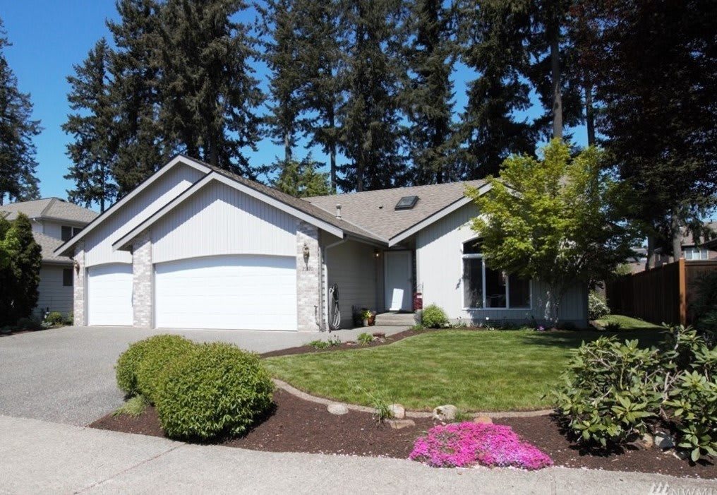 A suburban house with a well-maintained garden and driveway, surrounded by tall trees.