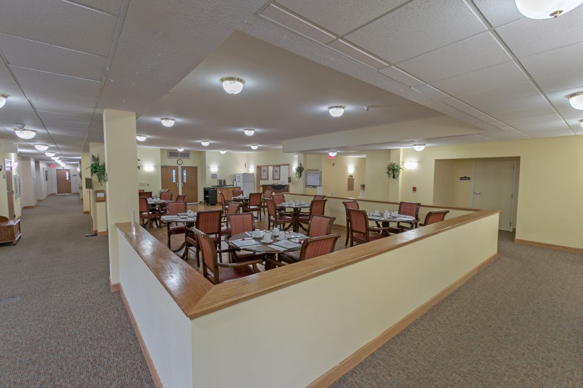 A spacious dining area with neatly arranged tables and chairs, featuring warm lighting and a carpeted floor.