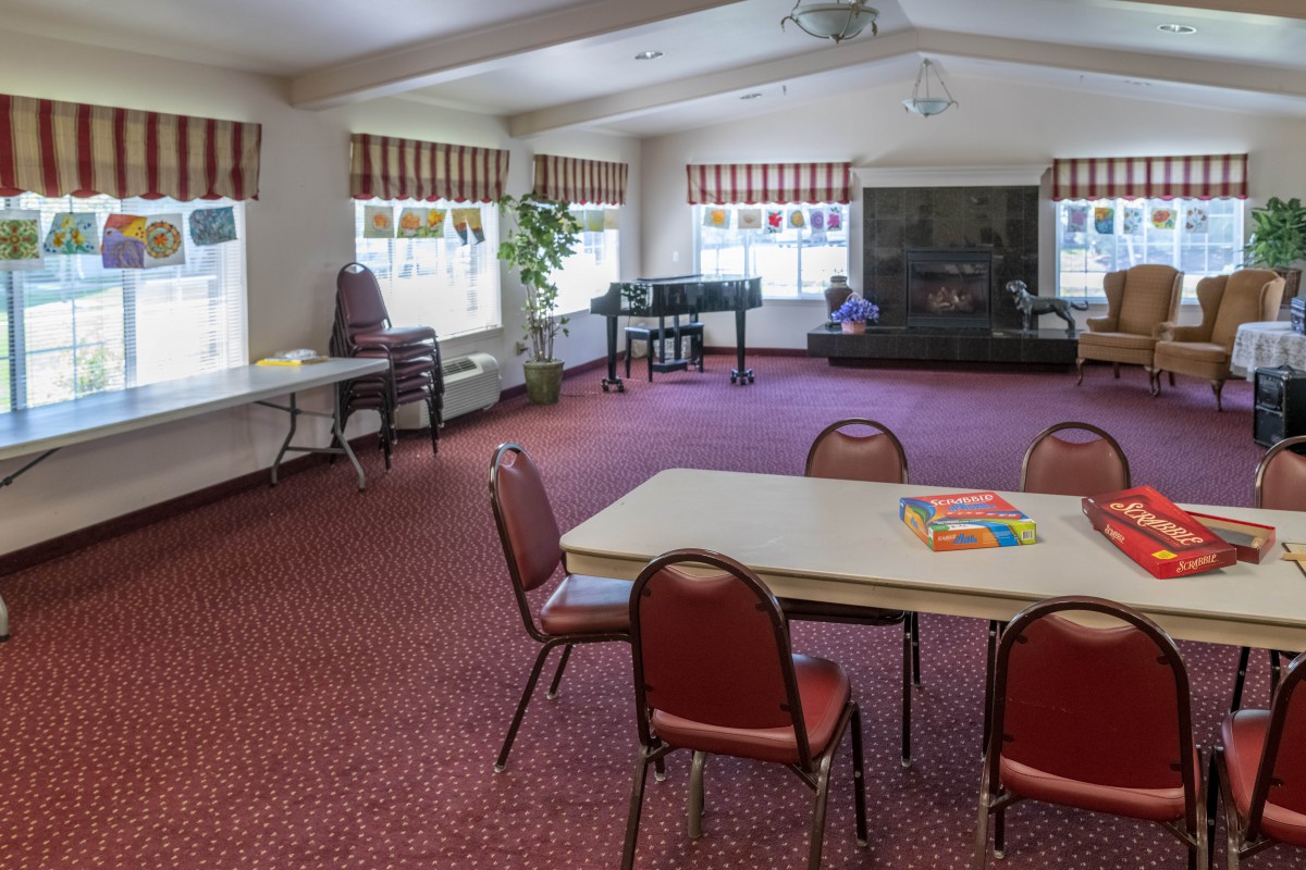 A spacious community room with tables and chairs, a piano, and large windows providing natural light.
