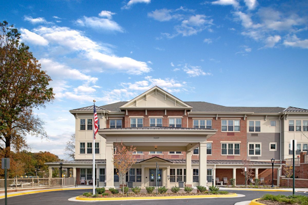 A three-story assisted living facility with a welcoming entrance and an American flag in front, set against a partly cloudy sky.