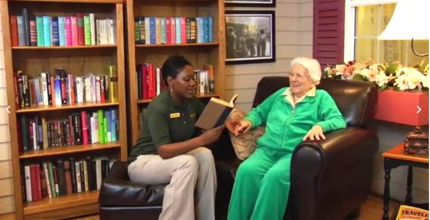 A caregiver reads a book to an elderly person in a cozy setting.