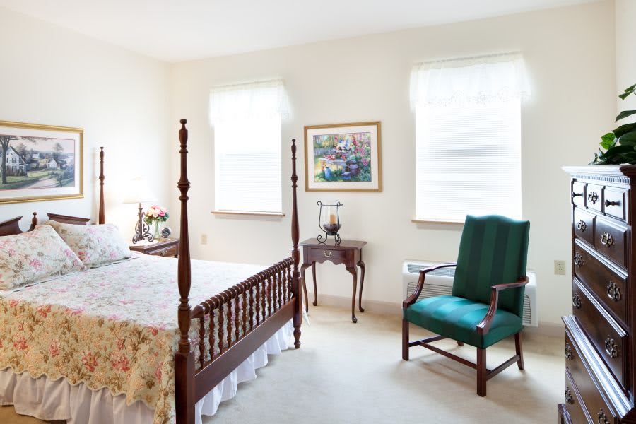A traditional bedroom featuring a wooden four-poster bed, a green upholstered chair, and classic decor with a painting on the wall.