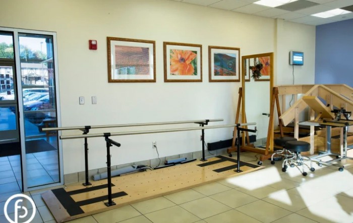 A rehabilitation room with parallel bars, a mirror, and framed artwork on the wall.