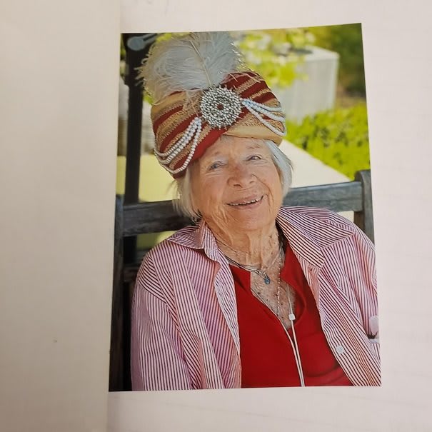 An elderly woman wearing a colorful hat sits outdoors, smiling warmly.