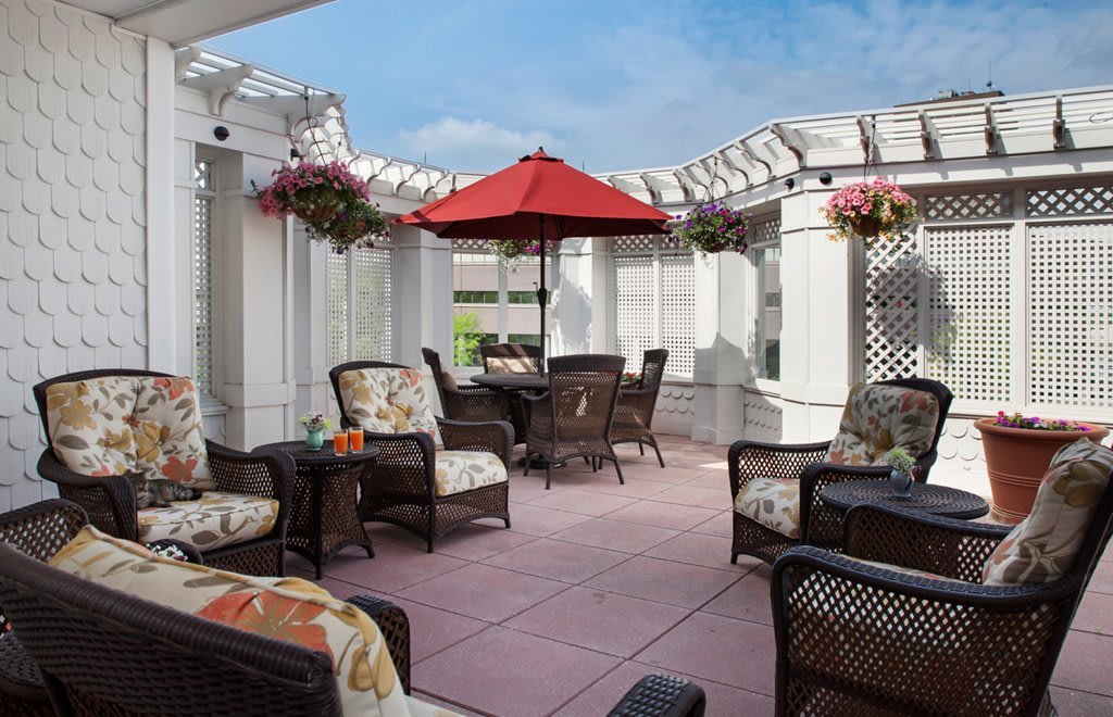A cozy outdoor patio with wicker seating, a red umbrella, and hanging plants under a pergola on a sunny day.