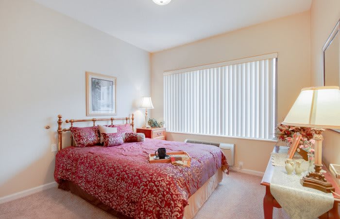 A cozy bedroom with a large bed covered in red bedding, illuminated by a table lamp next to a window with blinds.