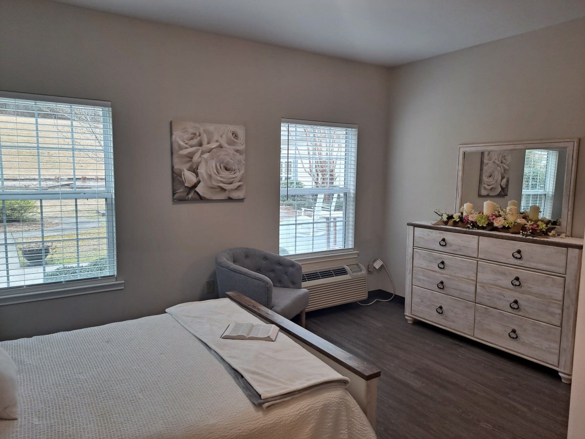 A cozy bedroom with a bed, upholstered chair, and a dresser with decorations under two windows.