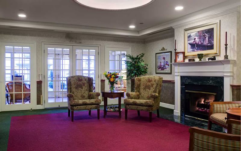 A cozy living room with two patterned armchairs, a small table adorned with flowers, and a fireplace surrounded by artwork.