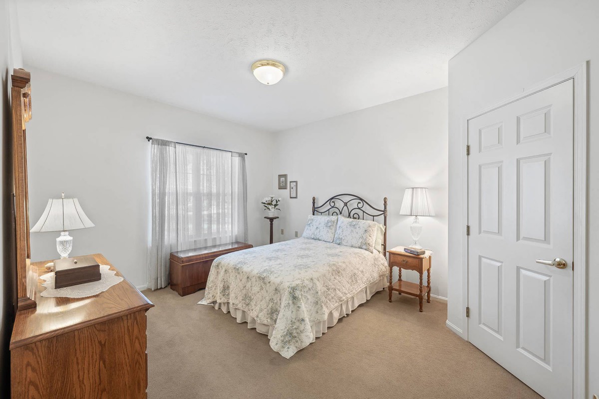 A cozy bedroom featuring a neatly made bed, a wooden dresser, and a bedside lamp next to a window with sheer curtains.