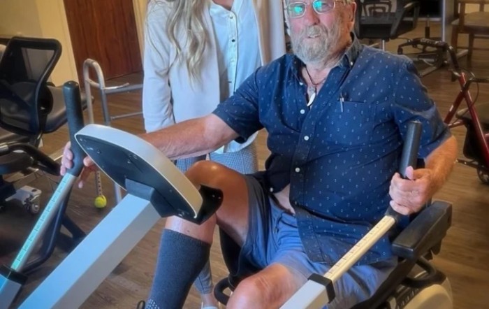 An elderly man using a stationary bike with a supportive person nearby.