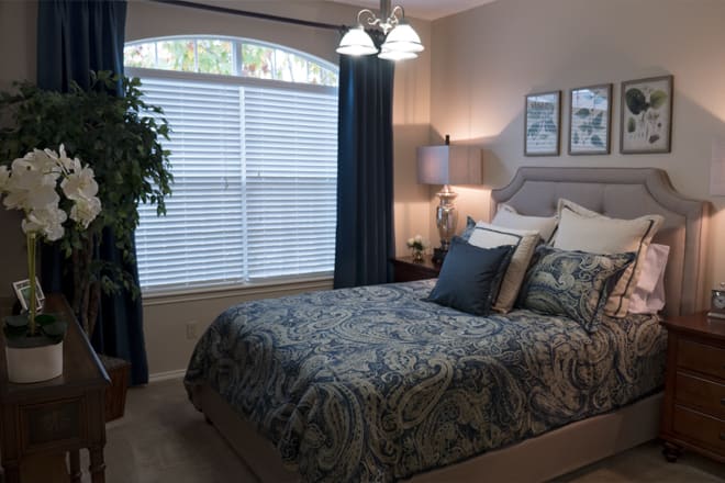 A cozy bedroom with a neatly made bed, decorative pillows, and soft lighting from a lamp. The room features a window with blinds and curtains, adding to the serene atmosphere.