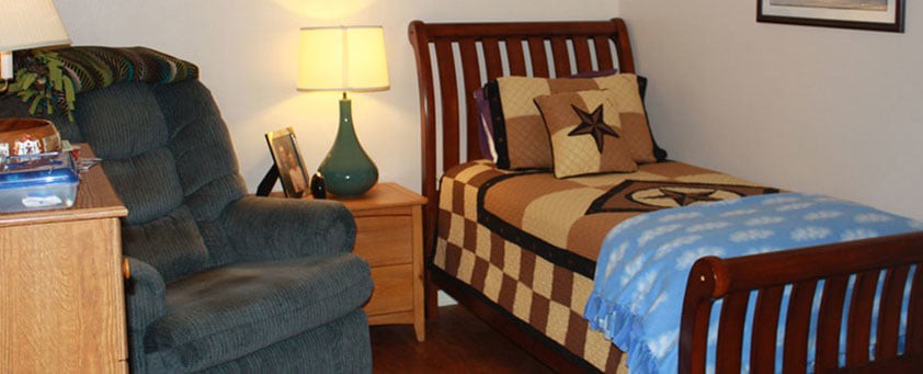 A cozy bedroom featuring a bed with a checkered quilt and star-themed pillows, accompanied by a nightstand with a lamp and chair.