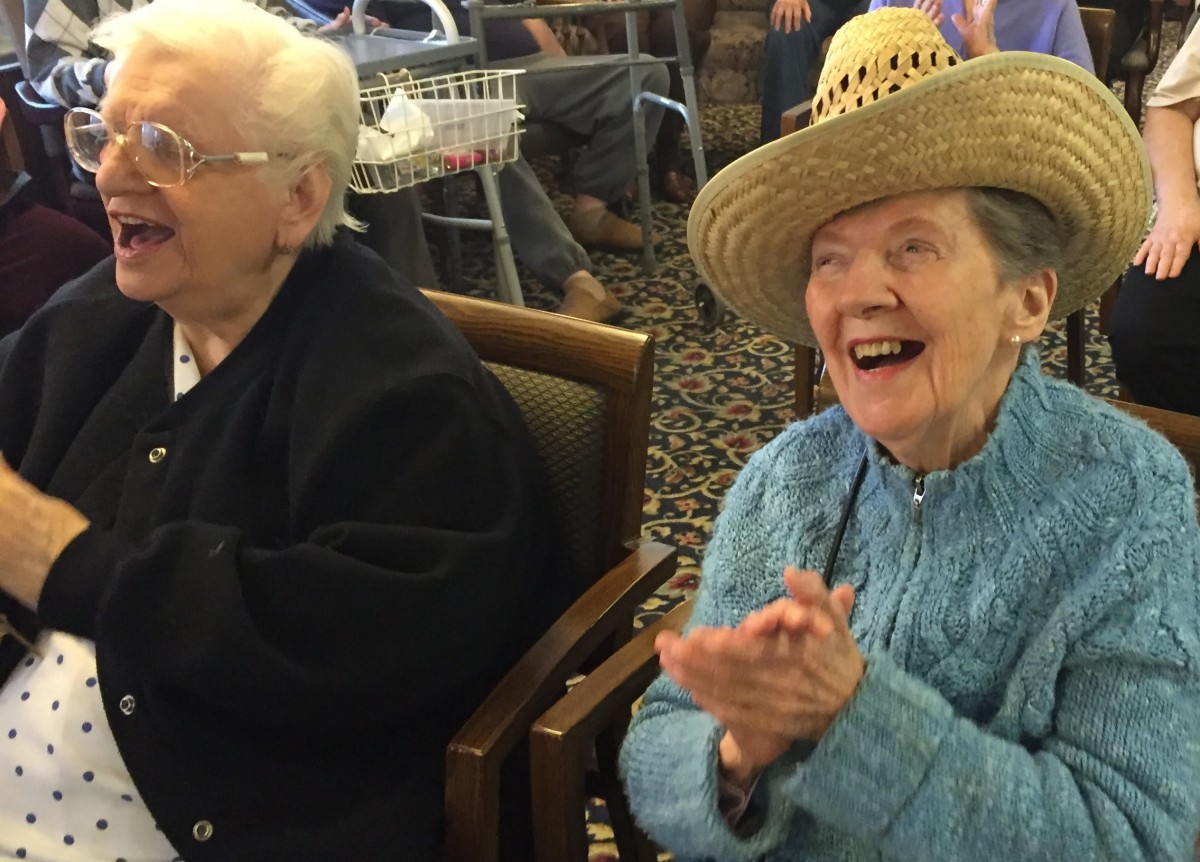 Elderly individuals enjoying a moment of laughter indoors, with one wearing a straw hat and a knitted sweater.