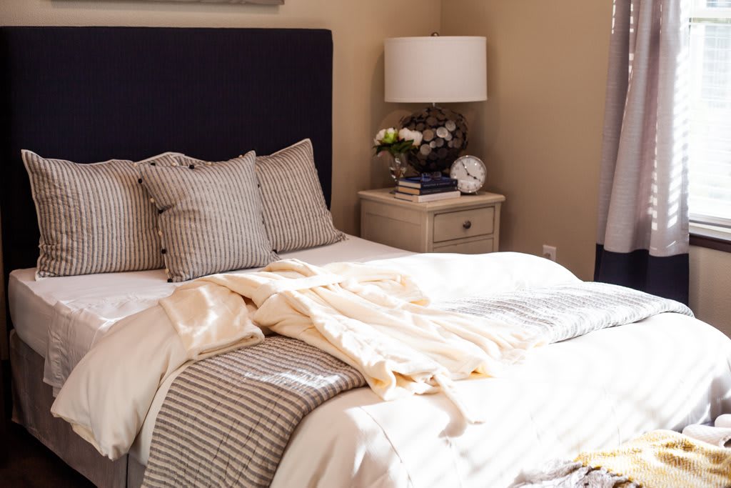 A cozy bedroom with a neatly made bed, pillows, and a nightstand featuring a lamp and decorative items.