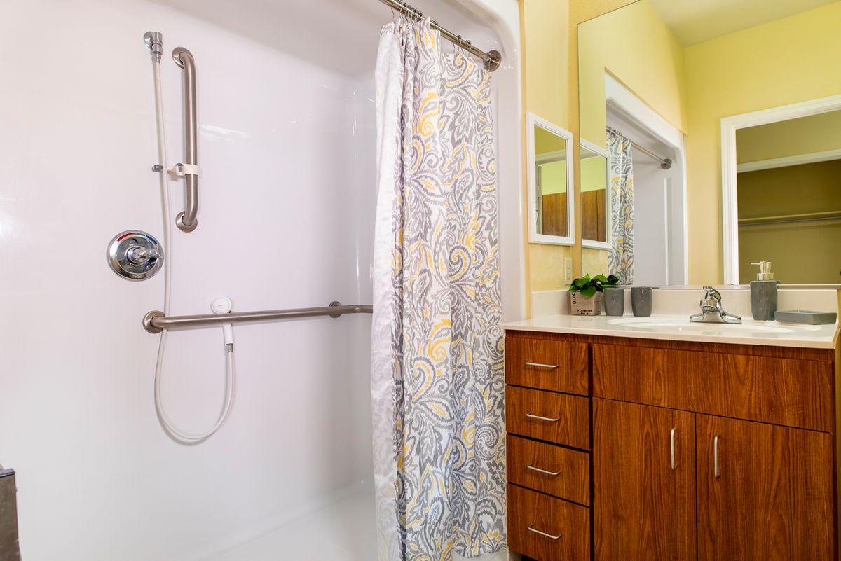 A modern bathroom featuring a shower with a grab bar and patterned curtain, adjacent to a vanity with wooden cabinetry.