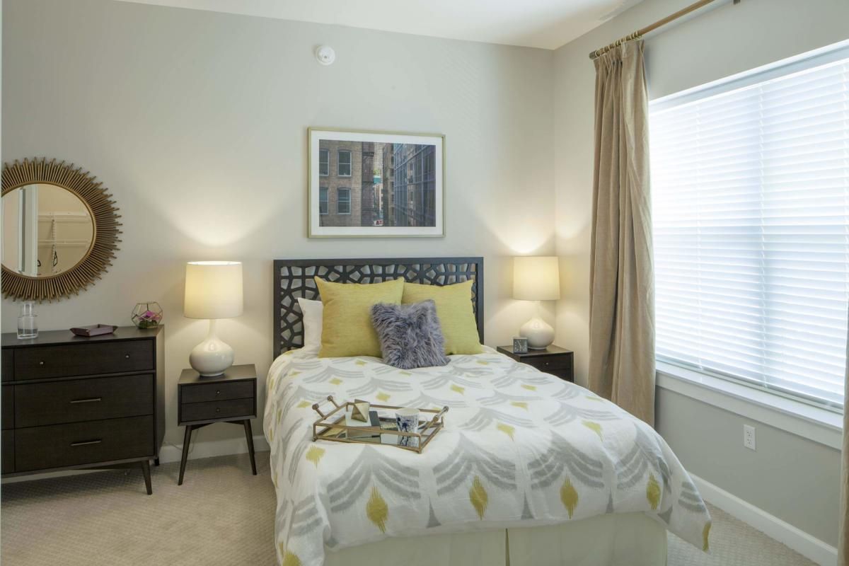A cozy bedroom with a neatly made bed, adorned with decorative pillows and flanked by two bedside lamps. Neutral-toned curtains frame the window, creating a serene atmosphere.